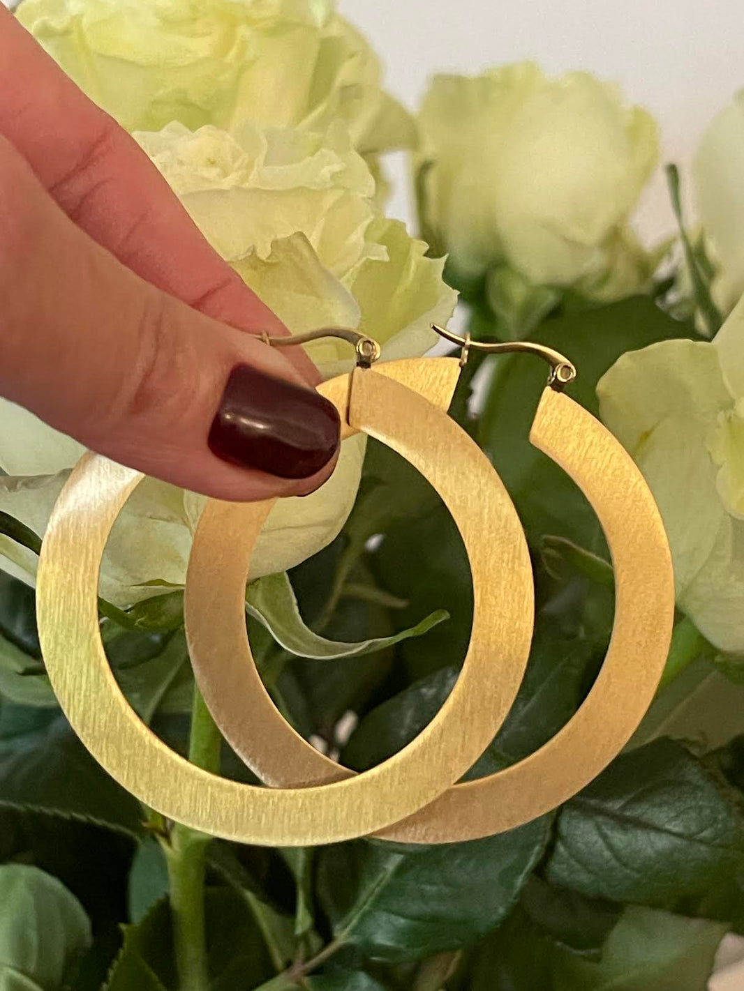 Mano sosteniendo dos argollas planas de acero inoxidable dorado, de 6 cm, frente a rosas blancas