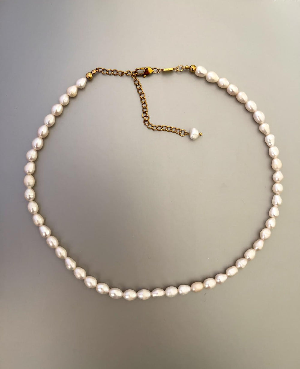 Collar corto de perlas de agua dulce blancas y ovaladas, con broche y cadena de alargue dorados, sobre fondo gris.