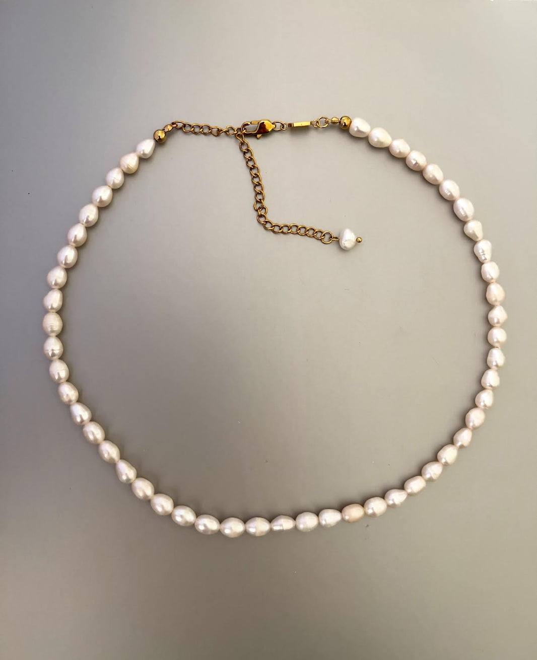 Collar corto de perlas de agua dulce blancas y ovaladas, con broche y cadena de alargue dorados, sobre fondo gris.
