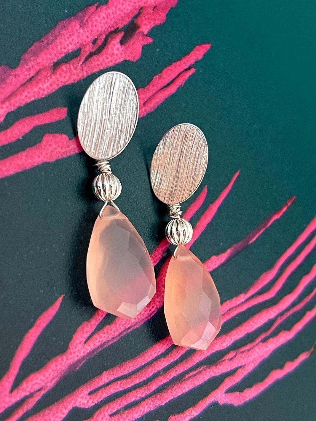Aros plateados con dos gotas grandes y facetadas de calcedonia rosa, sobre un fondo verde azulado con trazos rosados