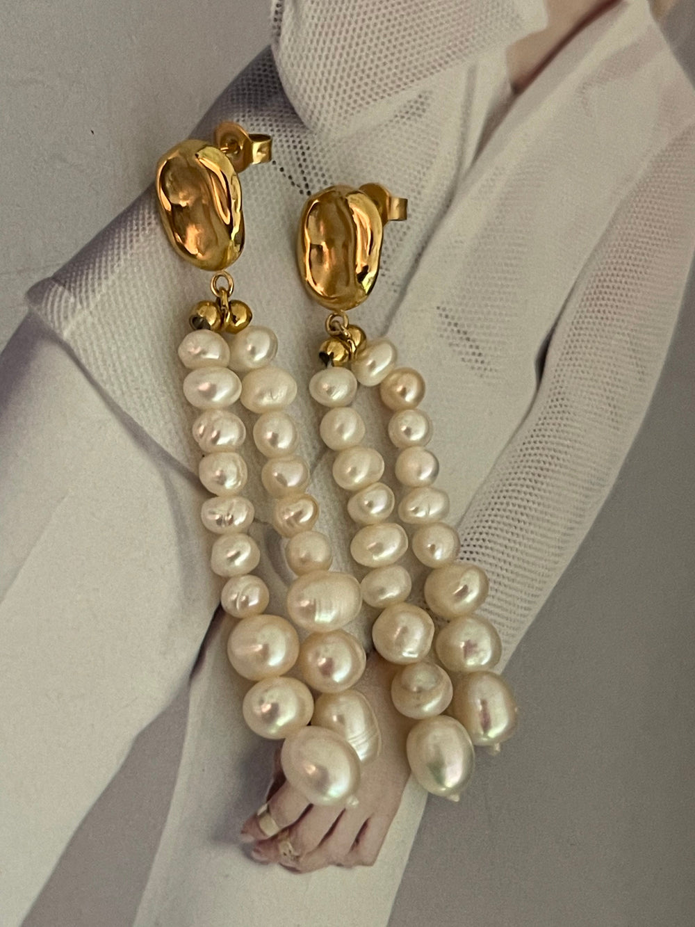 Aros con un botón dorado de forma irregular y tres hileras de perlas blancas que caen largas, sobre una tela clara.