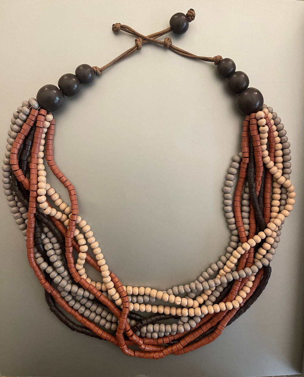 Collar largo de maderas en terracota, crema y gris, con cierre de cordón ajustable, sobre un fondo gris