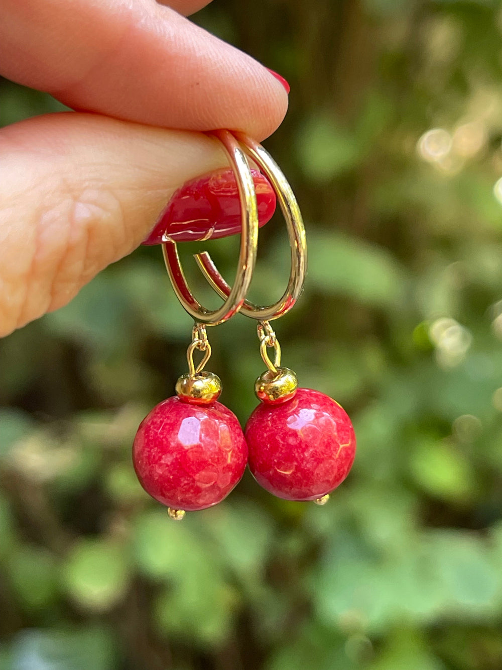 Dos aros de argolla dorada con una bolita roja de ágata, sostenidos con la mano frente a hojas verdes.